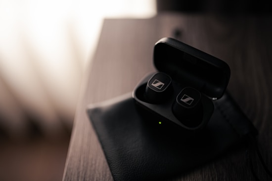 A pair of wireless earbuds rests inside an open charging case on a dark wooden surface. The case and earbuds have a sleek, black design with a small green light indicating power or connectivity status. Soft lighting creates a shadowed and moody atmosphere.
