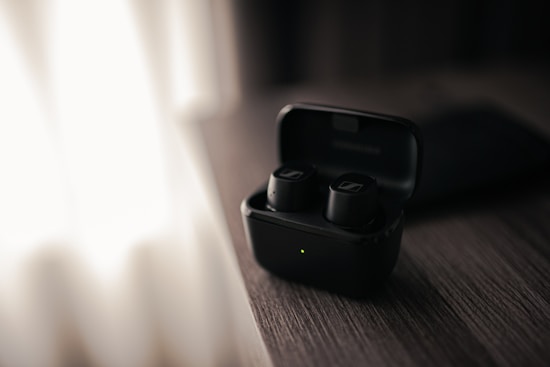 A pair of black wireless earbuds sit in an open charging case on a wooden surface. Soft, natural light from a nearby window creates a moody, calm atmosphere.