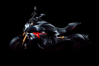 A sleek, modern motorcycle with a matte black finish is presented against a dark background. The motorcycle features notable elements such as bright orange suspension components and a distinctive red framework. The design highlights the cutting-edge engineering and aerodynamic structure, including a wide, comfortable seat and powerful-looking engine.