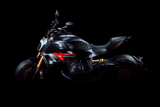 A sleek, modern motorcycle with a matte black finish is presented against a dark background. The motorcycle features notable elements such as bright orange suspension components and a distinctive red framework. The design highlights the cutting-edge engineering and aerodynamic structure, including a wide, comfortable seat and powerful-looking engine.