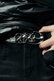 A close-up of a person wearing a stylish black leather belt with a polished, ornate metal buckle. The person is dressed in dark clothing, and their hand can be seen adjusting or holding the belt. The fabric of the clothing has a glossy finish, suggesting a high-fashion or edgy style.