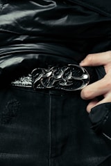 A close-up of a person wearing a stylish black leather belt with a polished, ornate metal buckle. The person is dressed in dark clothing, and their hand can be seen adjusting or holding the belt. The fabric of the clothing has a glossy finish, suggesting a high-fashion or edgy style.