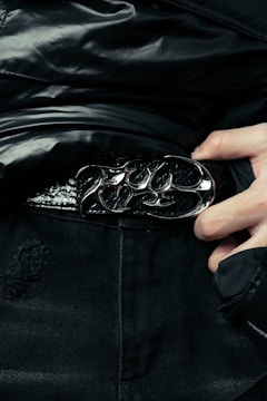 A close-up of a person wearing a stylish black leather belt with a polished, ornate metal buckle. The person is dressed in dark clothing, and their hand can be seen adjusting or holding the belt. The fabric of the clothing has a glossy finish, suggesting a high-fashion or edgy style.