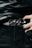 A close-up of a person wearing a stylish black leather belt with a polished, ornate metal buckle. The person is dressed in dark clothing, and their hand can be seen adjusting or holding the belt. The fabric of the clothing has a glossy finish, suggesting a high-fashion or edgy style.
