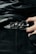 A close-up of a person wearing a stylish black leather belt with a polished, ornate metal buckle. The person is dressed in dark clothing, and their hand can be seen adjusting or holding the belt. The fabric of the clothing has a glossy finish, suggesting a high-fashion or edgy style.