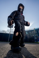 A person wearing a black puffer jacket, oversized black pants, and chunky black shoes stands outdoors against a clear blue sky. The jacket hood and face mask partially obscure their face. The camera angle is low, giving the person a larger-than-life appearance, and there are buildings and trees in the background.