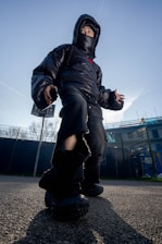 A person wearing a black puffer jacket, oversized black pants, and chunky black shoes stands outdoors against a clear blue sky. The jacket hood and face mask partially obscure their face. The camera angle is low, giving the person a larger-than-life appearance, and there are buildings and trees in the background.