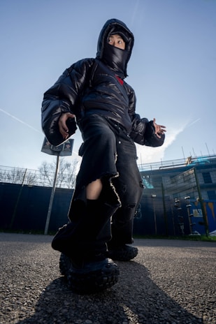 A person wearing a black puffer jacket, oversized black pants, and chunky black shoes stands outdoors against a clear blue sky. The jacket hood and face mask partially obscure their face. The camera angle is low, giving the person a larger-than-life appearance, and there are buildings and trees in the background.