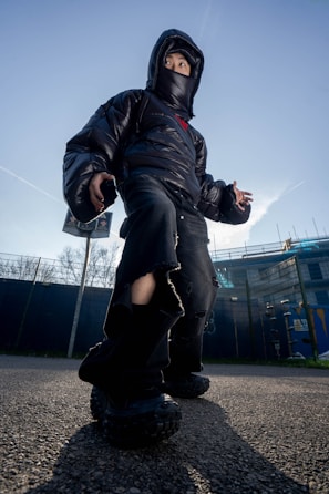 A person wearing a black puffer jacket, oversized black pants, and chunky black shoes stands outdoors against a clear blue sky. The jacket hood and face mask partially obscure their face. The camera angle is low, giving the person a larger-than-life appearance, and there are buildings and trees in the background.