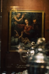 A framed painting is displayed behind a glass surface, depicting a scene with religious figures. The artwork features three adult figures and an infant, with expressions suggesting a moment of reverence or blessing. The surrounding setting has a soft, dark ambiance, and the glass reflects some light and objects from the room.