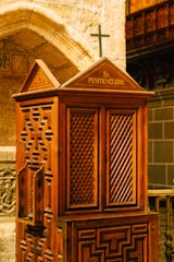 An intricately designed wooden confessional booth featuring lattice patterns and a small cross on top is situated within a stone-walled interior. The woodwork displays geometric carvings, and the setting suggests a religious or historical ambiance.