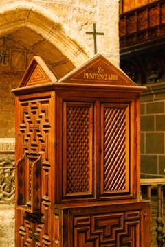 An intricately designed wooden confessional booth featuring lattice patterns and a small cross on top is situated within a stone-walled interior. The woodwork displays geometric carvings, and the setting suggests a religious or historical ambiance.