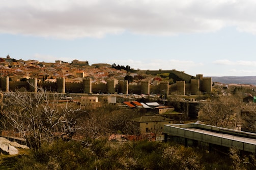 A historical cityscape featuring a sprawling town with a prominent ancient stone wall fortified with large round towers. The buildings display red-tiled roofs and are set against a hilly background. The foreground includes leafless trees and a modern structure, contrasting with the old architecture.