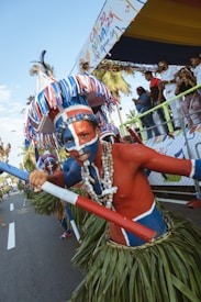 A person wearing vibrant, traditional festival attire with striking face paint in blue and red colors. They are holding a decorated pole and wearing a grass skirt and a large, colorful feathered headdress. Around the neck are several sets of chunky, white necklaces. In the background, there are people watching the event from behind a barrier, and a banner overhead indicating a carnival. Palm trees can be seen in the street setting.