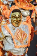 A person is dressed in a colorful, ornate costume featuring a shiny golden mask. The costume is decorated with bright orange and silver patterns, embellished with sequins and ruffles. The ensemble includes a detailed design that appears to be inspired by carnival or festival attire.