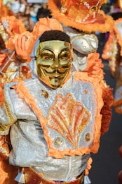 A person is dressed in a colorful, ornate costume featuring a shiny golden mask. The costume is decorated with bright orange and silver patterns, embellished with sequins and ruffles. The ensemble includes a detailed design that appears to be inspired by carnival or festival attire.