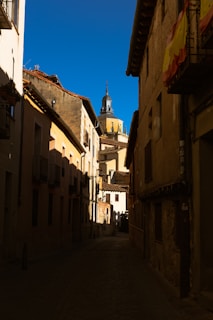 A narrow cobblestone street is lined with aged buildings, leading toward a towering dome and spire in the distance under a clear blue sky. Shadows from the late afternoon sun cast across the buildings, creating a dramatic contrast. A Spanish flag is draped from a balcony on the right.