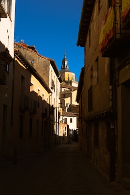 A narrow cobblestone street is lined with aged buildings, leading toward a towering dome and spire in the distance under a clear blue sky. Shadows from the late afternoon sun cast across the buildings, creating a dramatic contrast. A Spanish flag is draped from a balcony on the right.