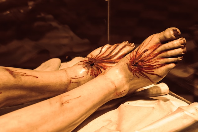A close-up view of two feet with wounds on top, displaying streaks of blood emanating from the injuries. The feet lie on a fabric surface in a dimly lit environment, possibly simulated or artificial in appearance.