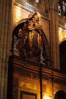 A richly decorated organ with gold detailing and intricate carvings is situated within a grand cathedral interior. Above the organ, there is a statue of an angel with wings spread, contributing to the ornate and reverent atmosphere. The surrounding architecture includes tall arches, stained glass windows, and stone walls. Natural light filters through the stained glass, casting colorful reflections.