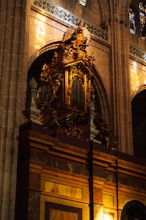 A richly decorated organ with gold detailing and intricate carvings is situated within a grand cathedral interior. Above the organ, there is a statue of an angel with wings spread, contributing to the ornate and reverent atmosphere. The surrounding architecture includes tall arches, stained glass windows, and stone walls. Natural light filters through the stained glass, casting colorful reflections.