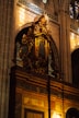 A richly decorated organ with gold detailing and intricate carvings is situated within a grand cathedral interior. Above the organ, there is a statue of an angel with wings spread, contributing to the ornate and reverent atmosphere. The surrounding architecture includes tall arches, stained glass windows, and stone walls. Natural light filters through the stained glass, casting colorful reflections.