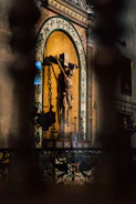 A religious statue of a crucifixion scene is set against an ornate wall with decorative patterns. The statue is surrounded by intricate ironwork and lit by soft, ambient lighting, casting dramatic shadows.