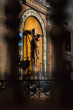 A religious statue of a crucifixion scene is set against an ornate wall with decorative patterns. The statue is surrounded by intricate ironwork and lit by soft, ambient lighting, casting dramatic shadows.