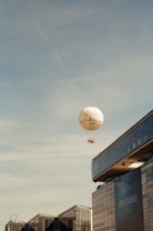 A hot air balloon is floating in a clear blue sky. Below, there are modern buildings with large glass windows, one of which features signage or advertisement.