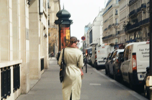 A woman wearing a beige trench coat walking through a softly lit urban street with neutral-toned buildings.
