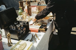 A table filled with various vintage and antique items is arranged outdoors. There are ornate glass decanters, brass objects, cutlery, and other trinkets. A person is reaching over the table, possibly examining or picking up an item.