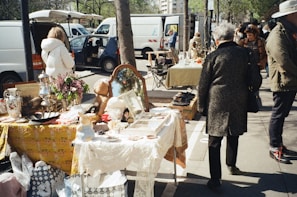 An outdoor market scene with tables set up along a sidewalk displaying various items for sale such as flowers, vases, a mirror, and other decorative objects. People walk by, browsing and interacting with the vendors under a sunny sky. White vans and trees line the background, adding to the bustling and lively atmosphere.