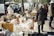 An outdoor market scene with tables set up along a sidewalk displaying various items for sale such as flowers, vases, a mirror, and other decorative objects. People walk by, browsing and interacting with the vendors under a sunny sky. White vans and trees line the background, adding to the bustling and lively atmosphere.