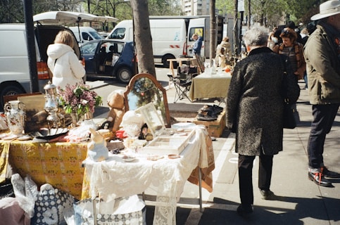 An outdoor market scene with tables set up along a sidewalk displaying various items for sale such as flowers, vases, a mirror, and other decorative objects. People walk by, browsing and interacting with the vendors under a sunny sky. White vans and trees line the background, adding to the bustling and lively atmosphere.