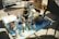A small table filled with various antique items, including lamps, a ceramic bust, glass bottles, a blue pitcher, and decorative figures. There is an assortment of other collectibles and household items scattered around. The setting appears to be indoors with natural light casting shadows on the floor and the table.