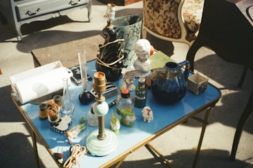 A small table filled with various antique items, including lamps, a ceramic bust, glass bottles, a blue pitcher, and decorative figures. There is an assortment of other collectibles and household items scattered around. The setting appears to be indoors with natural light casting shadows on the floor and the table.