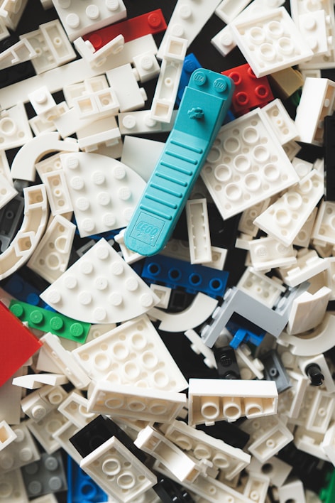 A scattered assortment of LEGO bricks in various shapes and colors, predominantly featuring white pieces with a standout teal brick among them. Other colors include red, black, blue, and green, with pieces in varying shapes such as rectangles and curves.