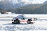 A sleek vehicle with a car body is modified with ski-like structures at the front and a track system at the rear, speeding across a snow-covered landscape. Mountains with snow caps and pine trees are visible in the background, suggesting a winter setting. The vehicle is emblazoned with racing decals and a slogan 'race to rescue'.