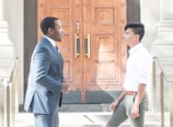 Two men are engaged in conversation in front of a large, intricately designed wooden door. One man is wearing a dark suit, while the other is dressed in a white shirt and green pants. They appear to be standing on stone steps with metal railings on either side.
