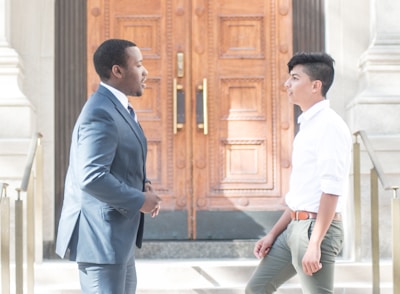 Two men are engaged in conversation in front of a large, intricately designed wooden door. One man is wearing a dark suit, while the other is dressed in a white shirt and green pants. They appear to be standing on stone steps with metal railings on either side.