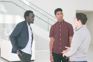 Three men are engaged in a conversation in a modern, well-lit setting. One man is wearing a navy blazer, another a maroon shirt, and the third a light-colored jacket. They stand near a stairway with glass and metal railings.