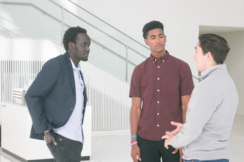 Three men are engaged in a conversation in a modern, well-lit setting. One man is wearing a navy blazer, another a maroon shirt, and the third a light-colored jacket. They stand near a stairway with glass and metal railings.