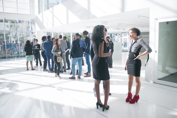 A group of people, mostly adults, are gathered in a modern, well-lit interior space with large windows. They appear to be engaged in conversation, standing in small groups. Two women are standing in the foreground, wearing stylish dresses and chatting with each other.
