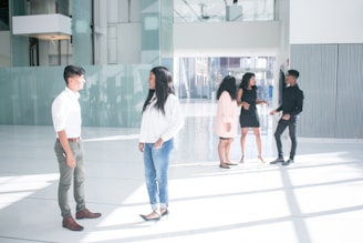A group of five individuals are standing in a spacious, modern interior space with large windows and abundant natural light. Two people are engaged in conversation in the foreground, while three others are interacting in the background. The setting appears to be an office lobby or atrium with a sleek design.