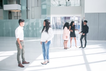 A group of five individuals are standing in a spacious, modern interior space with large windows and abundant natural light. Two people are engaged in conversation in the foreground, while three others are interacting in the background. The setting appears to be an office lobby or atrium with a sleek design.