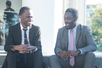 Two men sitting on a concrete ledge outside, dressed in formal business attire, and engaging in a lively conversation. They both are smiling and appear to be enjoying the moment.
