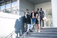 A group of young people are gathered outside a modern building, standing on stairs. They appear to be having a conversation, with some individuals smiling. The group includes both men and women dressed in formal and semi-formal attire, with one person holding a leather bag. The background features a glass entrance and a wall with text indicating an institution.