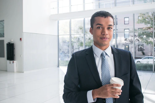 A person in a business suit stands in a well-lit modern office environment, holding a disposable coffee cup. Large windows in the background provide a view of the outdoors, where trees and buildings can be seen.