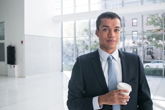 A person in a business suit stands in a well-lit modern office environment, holding a disposable coffee cup. Large windows in the background provide a view of the outdoors, where trees and buildings can be seen.