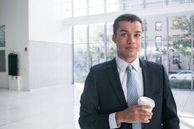 A person in a business suit stands in a well-lit modern office environment, holding a disposable coffee cup. Large windows in the background provide a view of the outdoors, where trees and buildings can be seen.